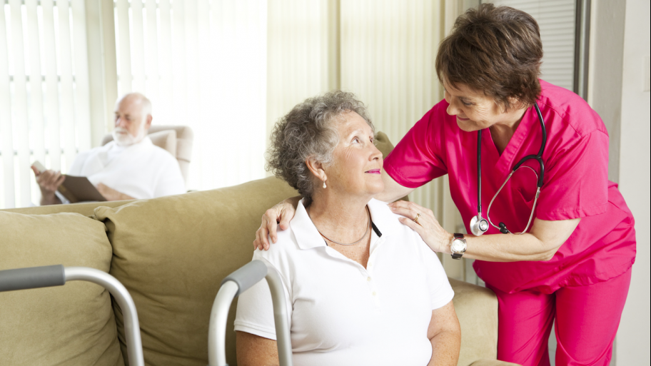 a medical professional talks with a woman in a nursing home facility who is seated on a coach with a walker nearby.